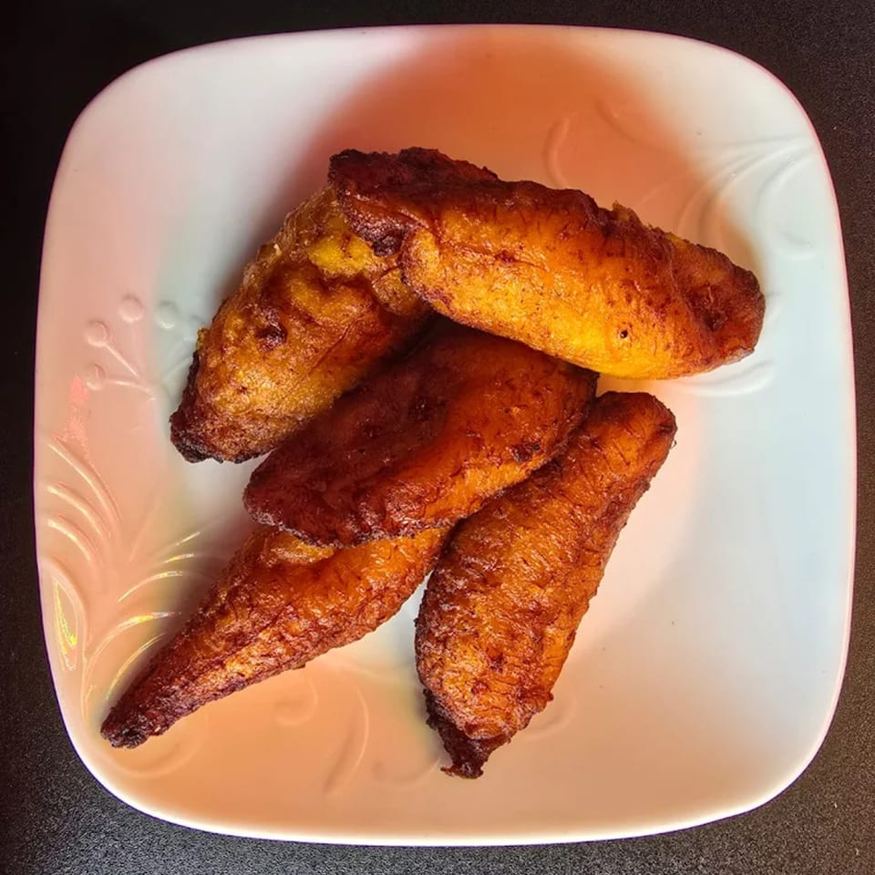 A white plate holds four pieces of golden-brown, fried Jamaican plantains, glistening with oil.
