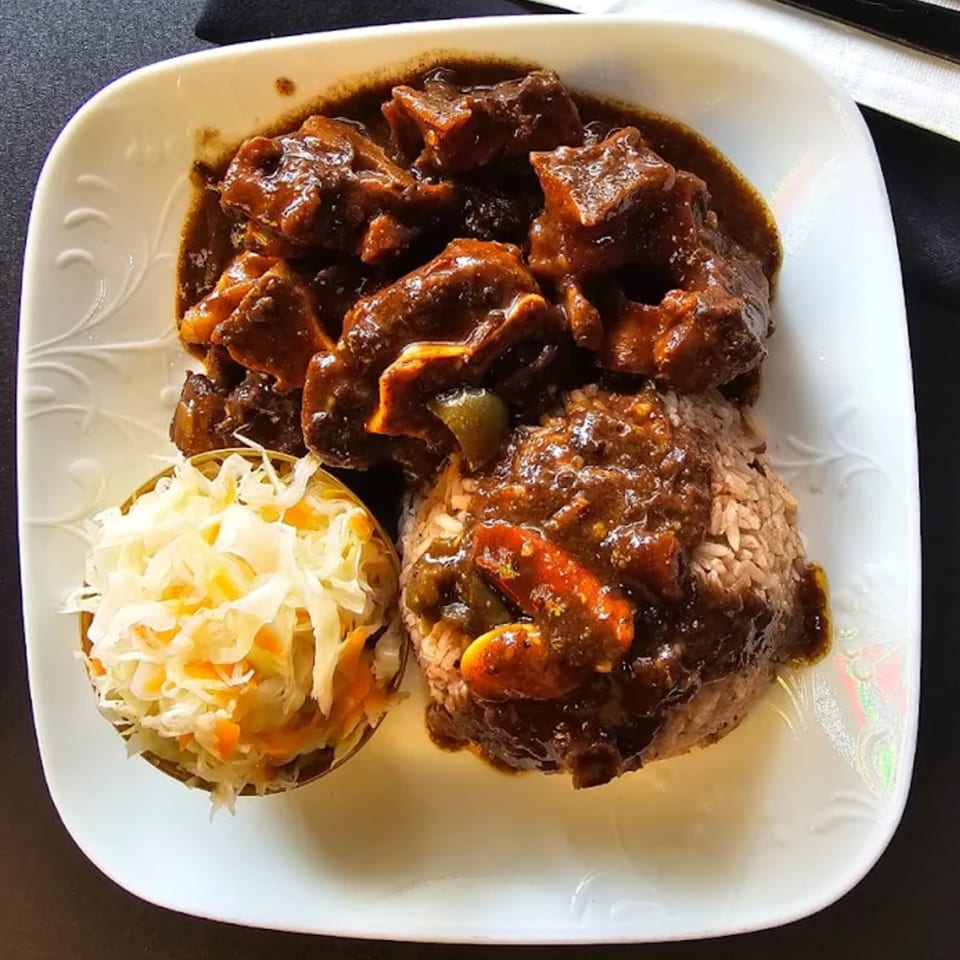 A plate of Jamaican oxtail stew with rice and peas, and a side of coleslaw.