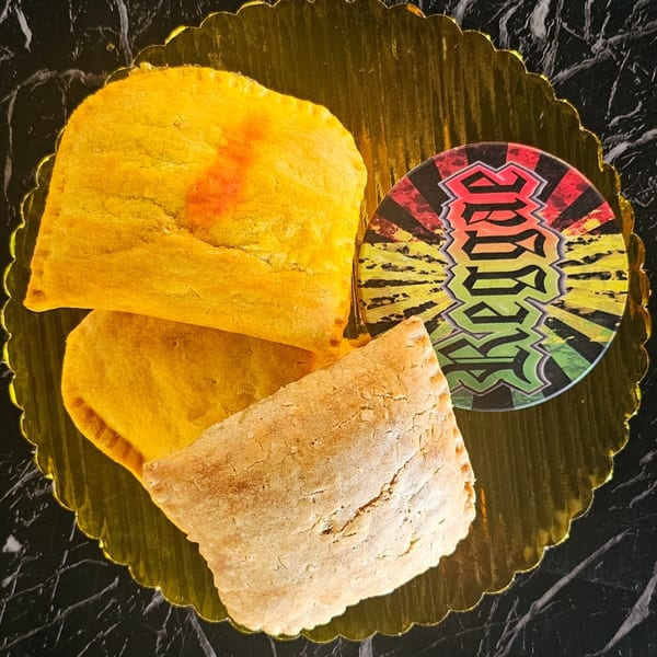 Three Jamaican patties, two golden and one light brown, on a gold scalloped tray with a Rasta-colored coaster.