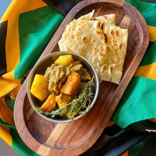 Jamaican curry goat with potatoes and roti bread on a wooden tray, with a Jamaican flag.