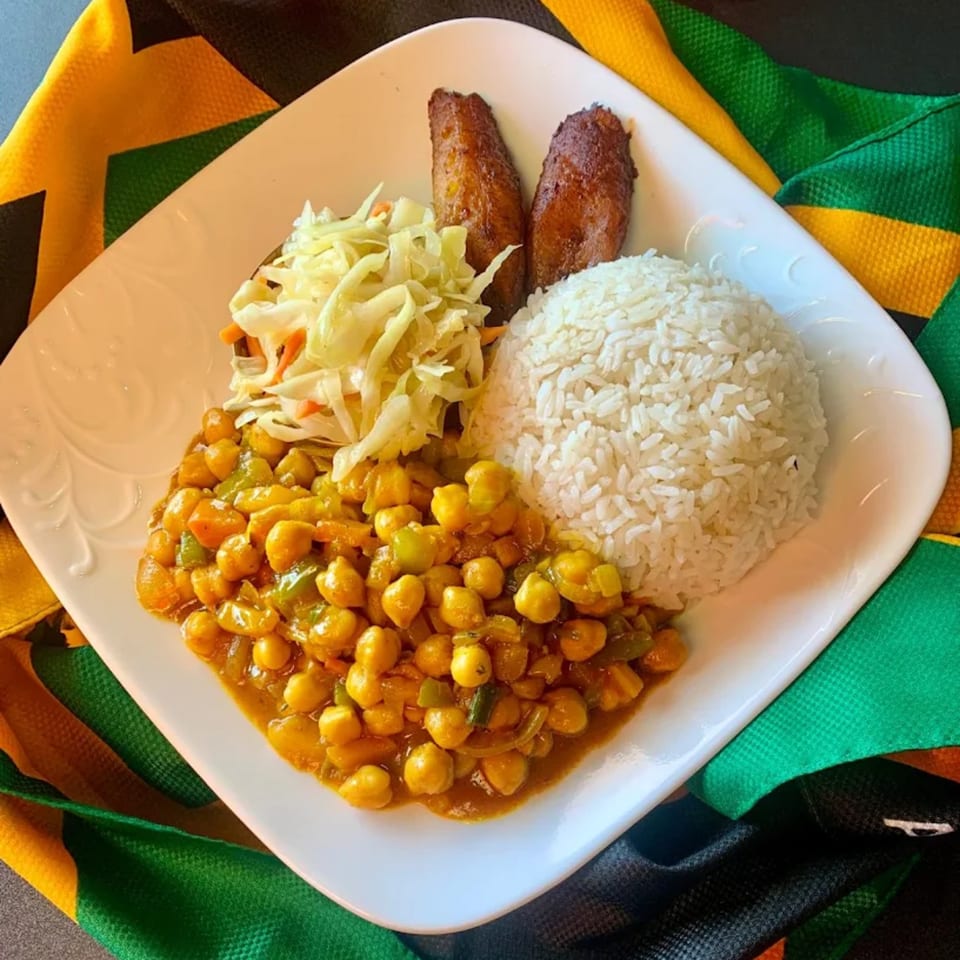 A Jamaican dish with curried chickpeas, white rice, coleslaw, and fried plantains on a white plate.