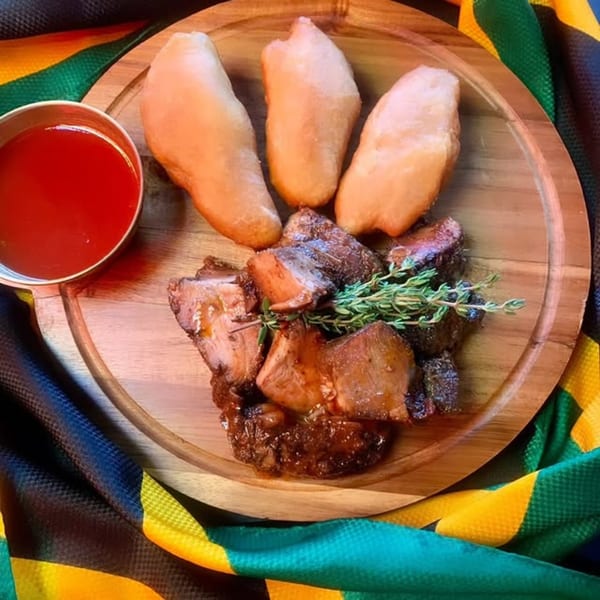 Jamaican stewed pork with three fried dumplings and a side of red sauce on a wooden plate.