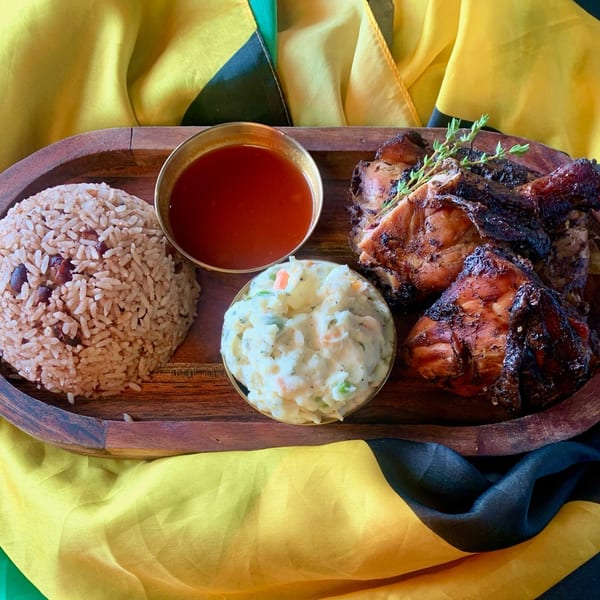 Jerk chicken with rice and peas, potato salad, and sauce on a wooden tray with a Jamaican flag.